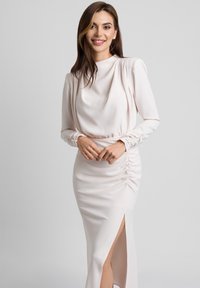 Jasnoróżowa sukienka z długim rękawem, z marszczonym materiałem, z bocznym rozcięciem i drapowanym dekoltem, uszyta z gładkiego, miękkiego materiału.