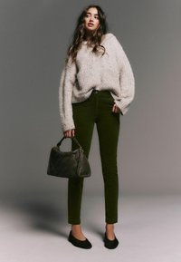 Pulover texturat de tricot gri deschis; pantaloni din corduroy verde închis; balerini negri; geantă matlasată de culoare închisă; model pe un fundal neutru.