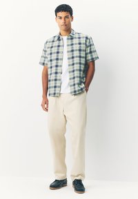 Short-sleeve plaid shirt in green, blue, and white, paired with cream trousers. Dark shoes with a chunky sole complete the outfit.