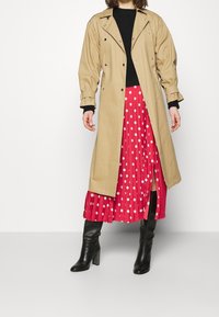 Imperméable beige avec des accessoires en métal argenté, superposé à une longue jupe plissée rouge à pois blancs. Des bottes noires jusqu'aux genoux complètent le look.