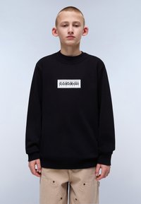 Svart hettegenser med ribbestrikket krage og mansjetter, med en hvit rektangulær logo på brystet, kombinert med lyse beige bukser.