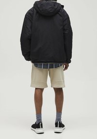 Czarna ocieplana kurtka z kapturem, noszona na koszuli w kratę, w połączeniu z beżowymi szortami i czarnymi sneakersami, widok z tyłu.