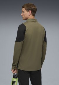 Olivenfarvet langærmet top med sorte skulderpaneler, tekstureret stof og krave. Modellen holder en grøn vandflaske.