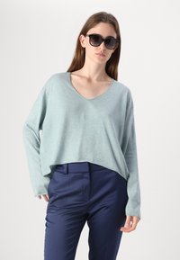 Světlomodrý zkrácený svetr s V-výstřihem a dlouhými rukávy, vyrobený z jemného materiálu, v kombinaci s tmavě modrými elegantními kalhotami a tmavými slunečními brýlemi.