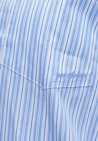 Sveto modro-belo tkanino z navpičnimi črtami krasi žep, na katerem je pri spodnjem desnem kotu nežno izvezen napis.