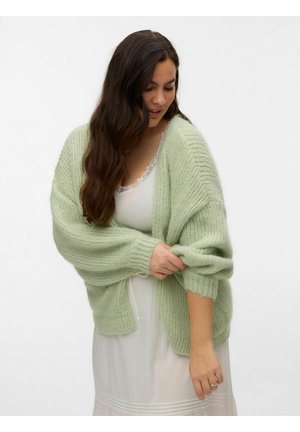 Frau mit langen dunklen Haaren, die eine hellgrüne grob gestrickte Strickjacke über einem weißen Spitzenkleid trägt und eine Ärmel hochkrempelt.