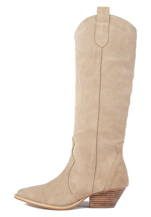 Bota de vaquero de ante beige con punta afilada, costuras decorativas y tacón de madera apilada. La altura llega a la mitad de la pantorrilla.
