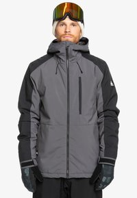 Veste imperméable grise avec accents noirs, fermeture �éclair à l'avant, capuche réglable et poche zippée. Le modèle porte des gants et des lunettes de ski.