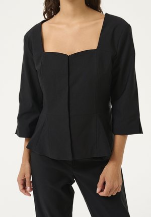 Vrouw draagt een aansluitende zwarte top met driekwart mouwen en een vierkante halslijn, gecombineerd met een zwarte broek, tegen een effen achtergrond.