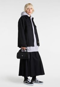 Black denim jacket over a gray hoodie, paired with a black maxi skirt. A small black bag with a handle and sneakers with white accents.