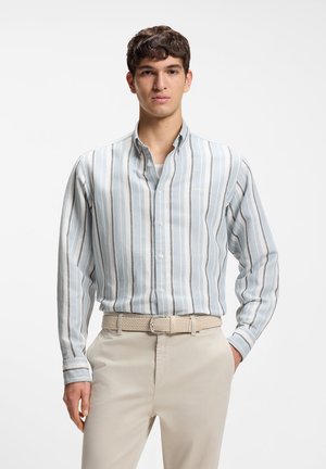 Νέος άνδρας που φοράει ένα ανοιχτό γαλάζιο και λευκό ριγέ πουκάμισο με κουμπιά, περασμένο μέσα σε μπεζ παντελόνι με πλεκτή ζώνη, στέκεται με το ένα χέρι στην τσέπη.