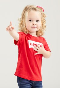 Rotes T-Shirt mit kurzen Ärmeln und weißem Levi's-Logo, kombiniert mit dunkelblauen Jeans. Kind mit lockigem blonden Haar und pinkem Haarband, das zeigt.