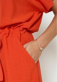 Orange, textured fabric dress with a relaxed fit and elastic waistband; features a side pocket and a silver bracelet on a hand.