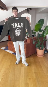 Tumehall hall sweater, millel on valge kirjas "YALE" ja bulldogi graafika. Modell hoiab seda üles. Toad on puidust põrandaga ja taustal on taimed.