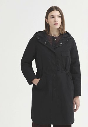 Jeune femme portant un manteau à capuche noir avec poches avant et boutons, debout de profil regardant vers la droite sur un fond uni.