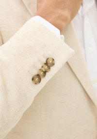 Beige linnen blazer mouw met vier ronde, lichtbruine knopen met vier gaten, en een wit overhemdmanchet die eronder zichtbaar is.