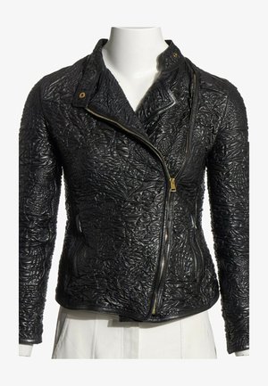 Chaqueta de cuero negro con patrones florales en relieve, cremallera asimétrica dorada y cuello con botón a presión, mostrada en torso de maniquí blanco.