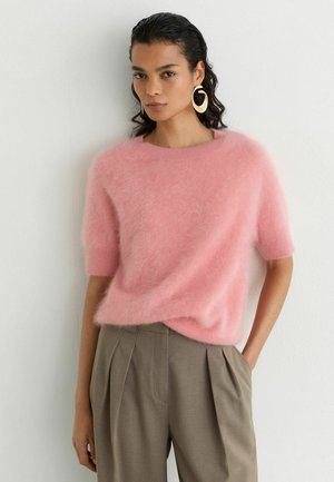 Vrouw met donker golvend haar draagt grote gouden oorringen, een roze zachte trui met korte mouwen en hoog getailleerde bruine geplooide broek.