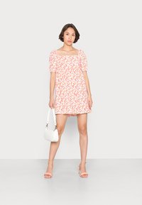 Vestido mini floral com mangas bufantes, decote quadrado, flores cor-de-rosa e amarelas sobre um fundo branco. A modelo carrega uma pequena bolsa branca.