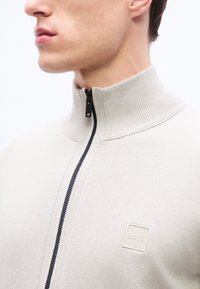 Κοντινό πλάνο ενός ατόμου που φοράει ανοιχτό μπεζ πουλόβερ με φερμουάρ και ψηλό γιακά, με ένα μικρό έμβλημα "BOSS" στο στήθος.