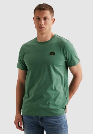 Man die een groene PME Legend T-shirt met korte mouwen en een blauwe spijkerbroek draagt, met één hand achter de rug staand tegen een effen achtergrond.