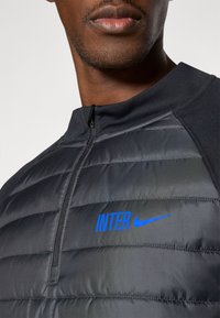 Jachetă neagră pentru bărbați, căptușită, cu fermoar, având textul „INTER” în albastru contrastant și logo-ul Nike, cu cusături în panouri și design cu guler înalt.