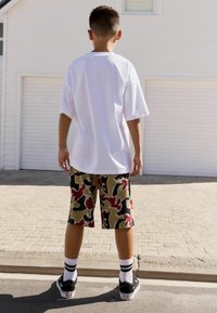 Witte oversized t-shirt, beige camouflaged shorts met rood en groen, zwarte sneakers en witte sokken met zwarte strepen.