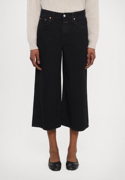Jean à jambe large et cropped noir en denim, avec une taille haute, cinq poches et des coutures contrastantes avec des accessoires de couleur cuivrée.