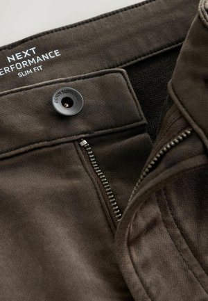 Mørkebrune slim fit jeans med lynlås delvist åbent, sort knap mærket "DENIM SUPPLY CO," og hvid trykt branding indvendigt i livlinjen.