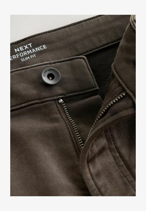 Jeans slim fit de culoare maro închis, cu fermoar parțial deschis, nasture negru cu eticheta "DENIM SUPPLY CO" și branding alb imprimat pe interiorul taliei.