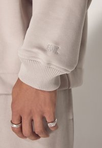 Manga de sudadera gris claro con puño acanalado, que presenta un discreto bordado de "EDIT." Mano visible adornada con anillos de plata.