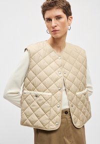 Colete bege acolchoado com botões de pressão, apresenta dois bolsos frontais e decote arredondado. Usado por cima de uma camisa de manga comprida creme.