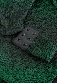 Zaļš teksturēts audums ar krokotu dizainu. Melna akcenta daļa ar sešām apaļām pogām līdzīgām detaļām. Mīksts materiāls ar nelielu spīdumu.