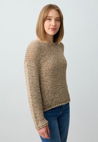 Beige gestrickter Pullover mit langen Ärmeln, rundem Ausschnitt und strukturierter Oberfläche. Kombiniert mit blauen Jeans, die einen lässigen Schnitt zeigen.