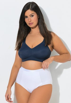 Žena s dlouhými tmavými vlasy, oblečená v tmavě modré sportovní podprsence a bílém spodním prádle s vysokým pasem, stojící na jednobarevném bílém pozadí.