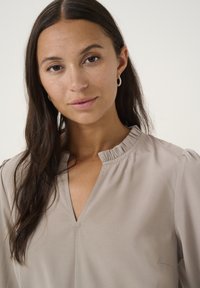 Beige halenka s volánovým límcem, výstřihem do V a měkkou tkaninou. Obsahuje jemnou texturu a kulaté rukávy.