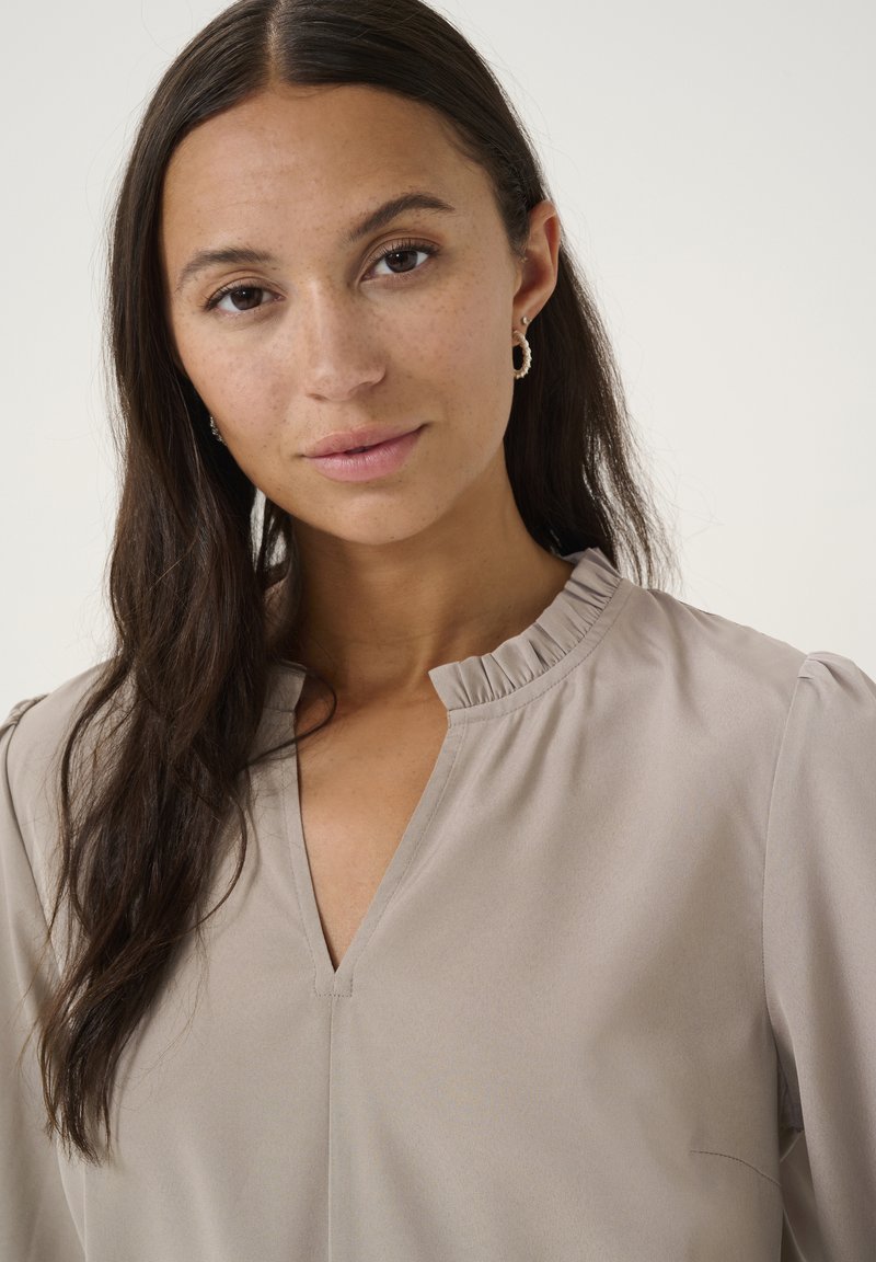 Beige halenka s volánovým límcem, výstřihem do V a měkkou tkaninou. Obsahuje jemnou texturu a kulaté rukávy.