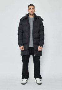 Doudoune noire avec capuche, dotée d'une fermeture éclair, de plusieurs poches et d'un design matelassé. Portée par-dessus un pull gris et un pantalon noir.
