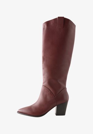 Bota alta hasta la rodilla de cuero burdeos con punta puntiaguda, tacón ancho y un detalle de lazo en la parte superior para facilitar su puesta. Acabado liso.