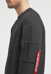 Sudadera negra con cuello acanalado, que incluye un bolsillo en la manga izquierda y una etiqueta roja con el texto "REMOVE BEFORE FLIGHT".