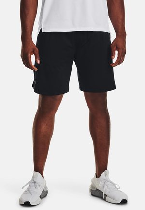 Homme portant un short de sport noir, des baskets blanches et une chemise blanche, debout devant un fond blanc uni.