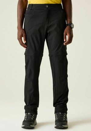 Ein Mann trägt schwarze Zip-off-Wanderhose, schwarze Wanderschuhe und ein gelbes Shirt und steht vor einem schlichten hellen Hintergrund.