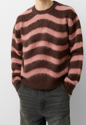 Mand iført en blød sweater med brede, bølgede vandrette striber i lyserød og mørkebrun, kombineret med falmede sorte jeans.