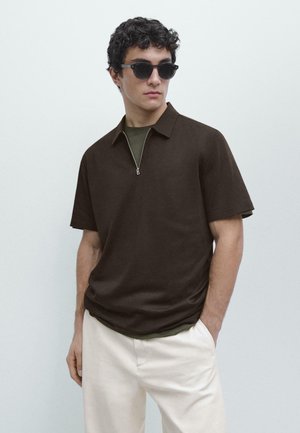 Giovane uomo con polo nera a maniche corte, pantaloni beige e occhiali da sole scuri, in piedi contro uno sfondo chiaro e uniforme.