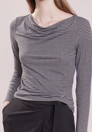 Woman wearing a black and white patterned long-sleeve top with a draped neckline and black pants with a tied waist detail.