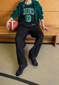 Pantaloni da basket neri, una maglia dei Celtics con scritte verdi e scarpe sportive viola e nere. Una palla da basket è poggiata sulla mano del soggetto.