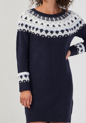 Femme portant une robe pull en tricot bleu marine avec des motifs géométriques blancs et bleu clair sur les épaules et les poignets.