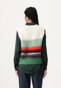 Sweter bez rękawów z białym tłem i poziomymi paskami w kolorach zielonym, czerwonym, czarnym i granatowym. Miękka struktura o luźnym kroju, nałożony na ciemną koszulę.