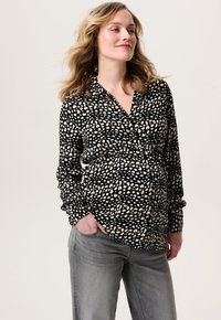 Zwarte blouse met een wit stippenpatroon, lange mouwen, knoopsluiting bij de kraag en een relaxte pasvorm, gedragen met losse grijze jeans. Zachte stofstructuur.