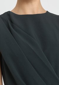 Ciemnozielony top bez rękawów z drapowanym wzorem, gładką fakturą tkaniny i szerokim dekoltem. Brak widocznych wzorów ani elementów metalowych.